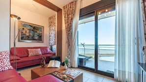 Uma sala de estar com um sofá vermelho, uma mesa de centro, um abajur, um quadro e vista para o mar através de uma porta de vidro deslizante.