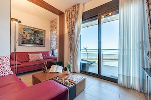 A living room with a red sofa, a coffee table, a floor lamp, a painting, and a view of the sea through a sliding glass door.