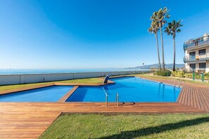 A swimming pool with a wooden deck, surrounded by grass and palm trees, with a view of the sea and mountains in the background. - Seaside Luxury Solarium Apartment with Sea Views, Wi-Fi, and Air Conditioning (Mont-roig Bahia)