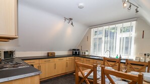 Une cuisine avec des meubles en bois, un plan de travail noir, un four à micro-ondes, une bouilloire et une fenêtre avec des stores.