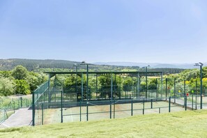 A tennis court with a net, surrounded by a metal fence, and a viewing platform with a clear view of the surrounding landscape.