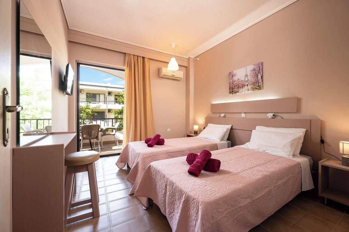 Habitación de hotel con dos camas, un escritorio, una silla, un televisor y un balcón con vistas a un edificio y árboles.