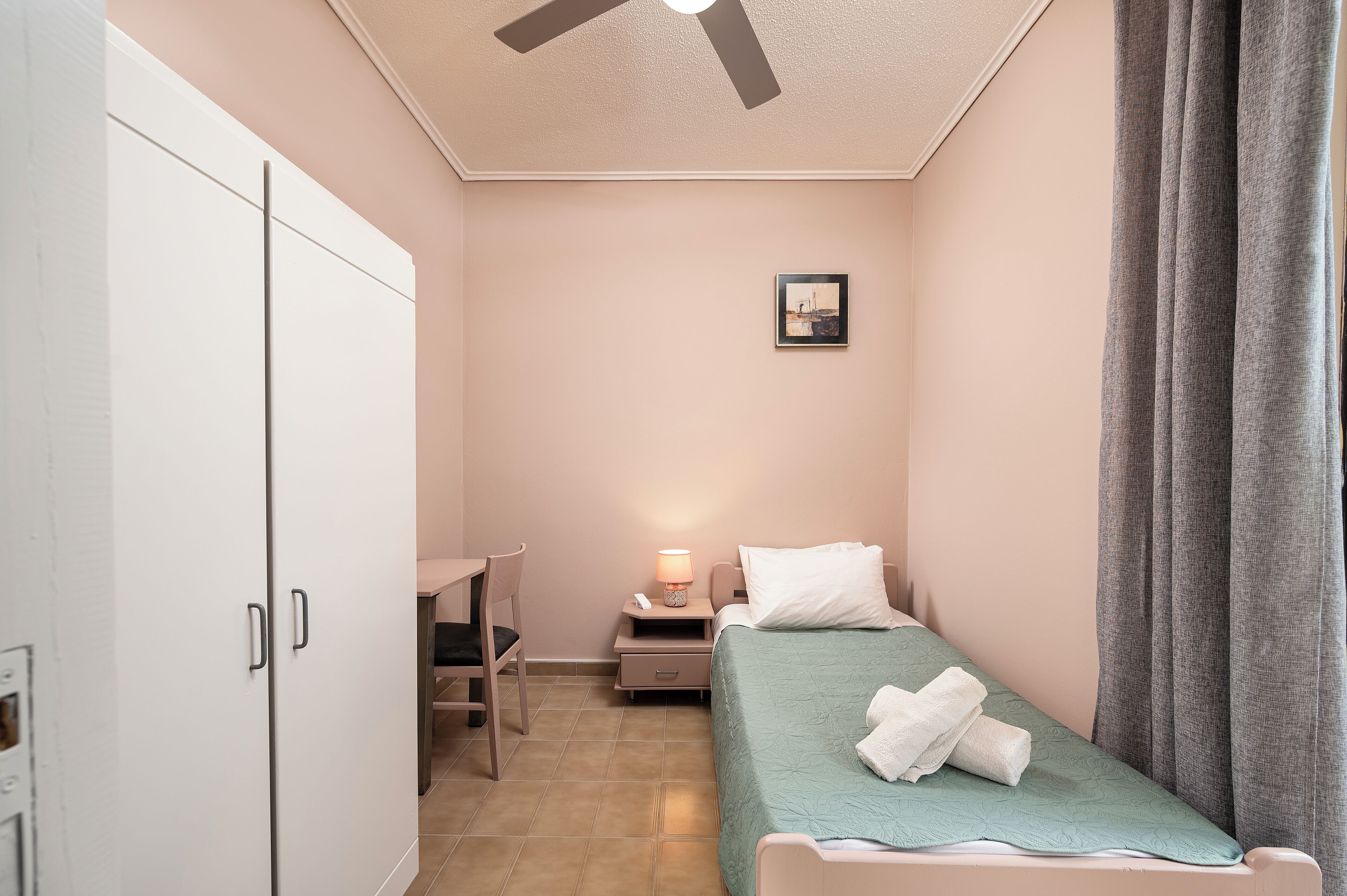 Quarto pequeno e bem iluminado com uma cama de solteiro, uma escrivaninha e um guarda-roupa.