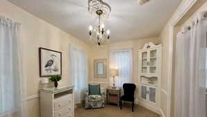 A well-lit room with a chandelier, a framed bird painting, a wicker console table, a patterned armchair, a black chair, and a cabinet with glass doors.