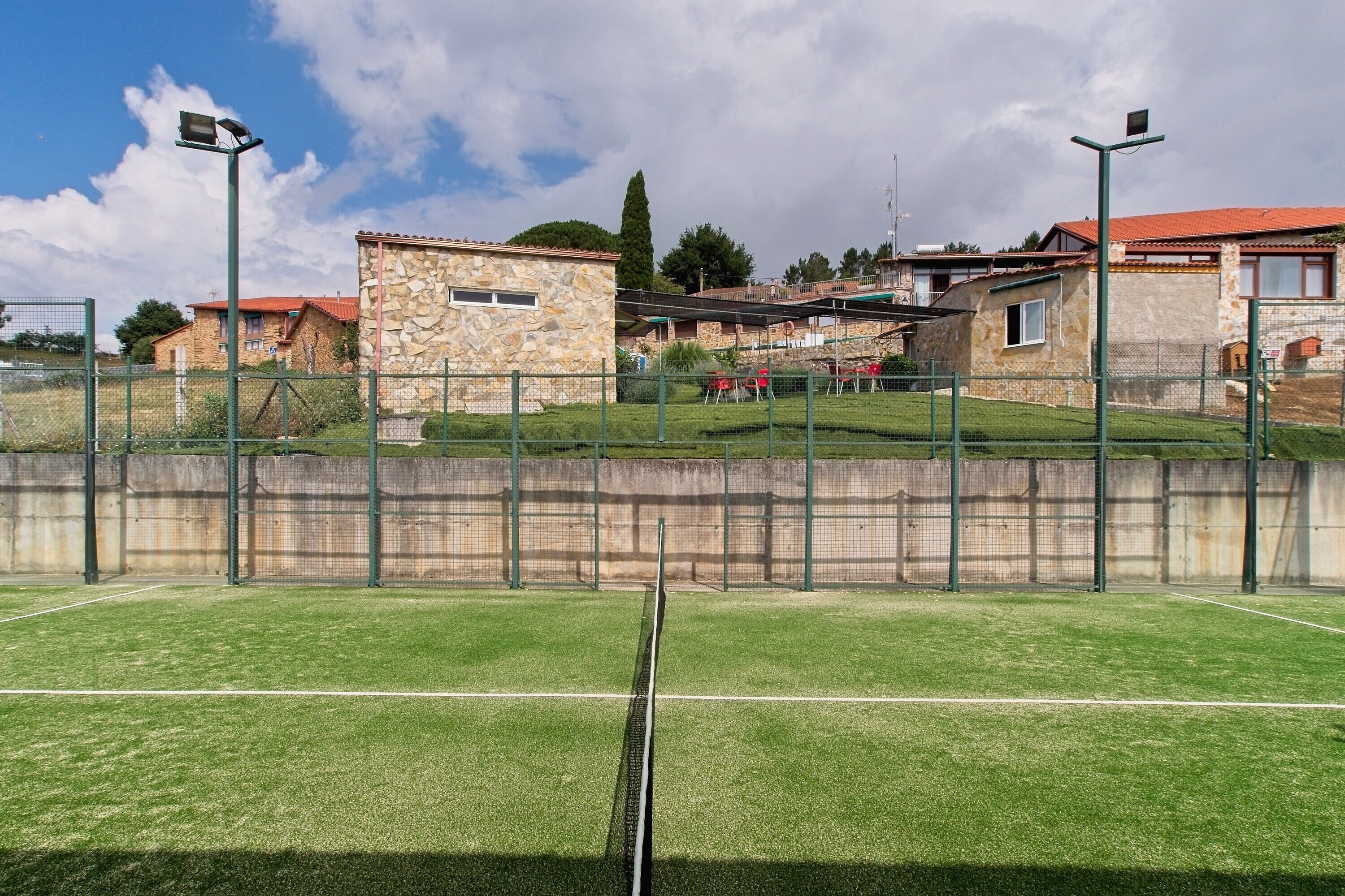 Een tennisbaan met een net, omgeven door een groene hekken, en huizen op de achtergrond.