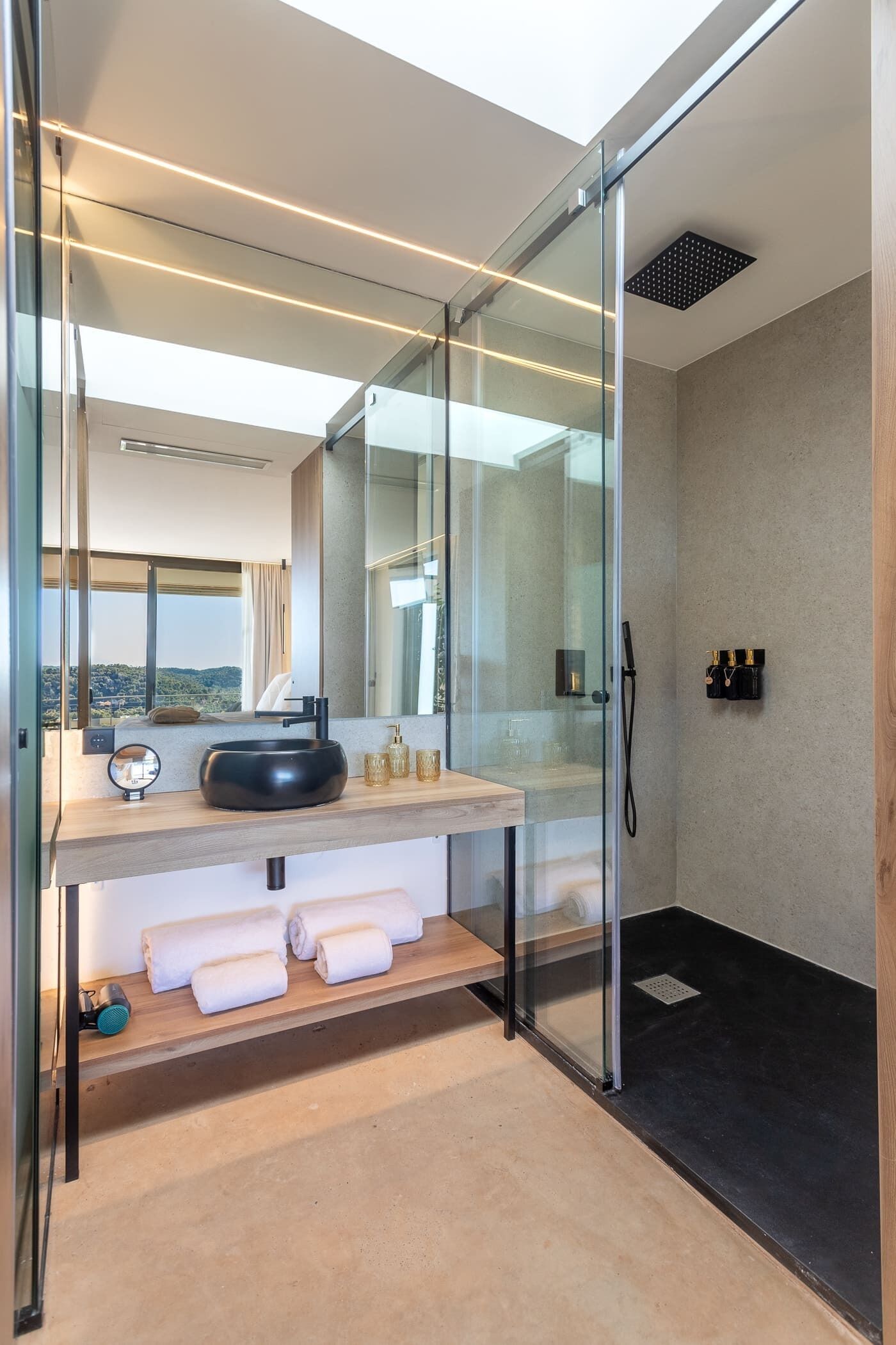 Un baño moderno con cabina de ducha de vidrio, un espejo redondo, un lavamanos negro y un tocador de madera.