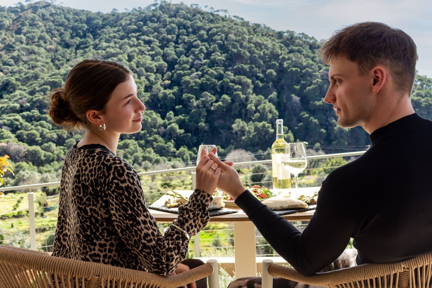 A couple dining outdoors with a table set for two, overlooking a forested hillside.