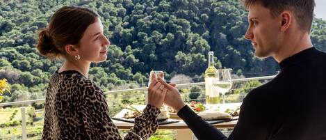 A couple dining outdoors with a table set for two, overlooking a forested hillside.