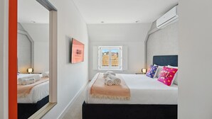 A modern hotel room with a large bed, a window with a view of buildings, a mirror, and a framed sign on the wall.