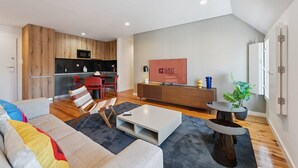 A modern living room with a sofa, coffee table, and a TV. There is a kitchen area with wooden cabinets and a microwave.