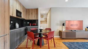 A modern kitchen with wooden cabinets, a black countertop, and a red toaster. There is a dining area with a round table and red chairs. A flat-screen TV is on a stand to the right.