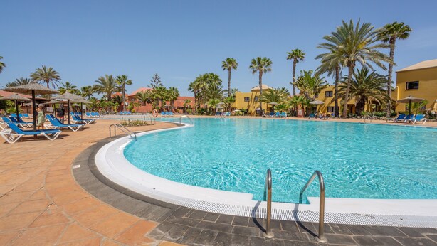 A swimming pool with blue lounge chairs, palm trees, and a clear sky. - Vacation 'Vista Lobos' with Sea View, Shared Pool and Wi-Fi (Corralejo)