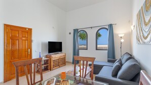 A living room with a television, wooden furniture, a sofa, and a view of the beach through two windows. - Vacation 'Vista Lobos' with Sea View, Shared Pool and Wi-Fi (Corralejo)