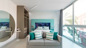 A modern bedroom with a large bed, a sofa, a small table with drinks, and a large window with a view of the street.