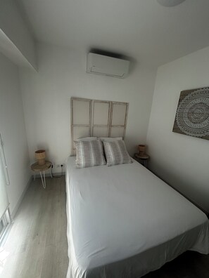 A bedroom with a bed, a bedside table, a wall-mounted air conditioner, a small wooden side table, and a decorative wall piece. - Charming, two-room apartment 500 m from the beach and all shops (Villeneuve-Loubet)