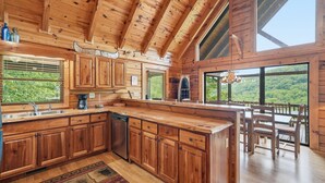 A wooden cabin with a kitchen featuring a large island, wooden cabinets, and a dining area with a view of the forest.