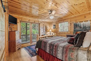 Quarto de cabana de madeira com cama, escrivaninha, televisão e ventoinha no teto.