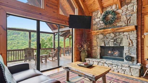 A wooden cabin interior with a stone fireplace, a flat-screen TV, and a balcony with a view of the forest.