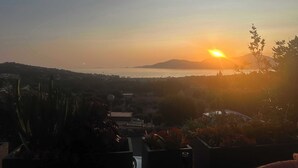 Une vue sur le coucher de soleil depuis un balcon donnant sur une étendue d’eau et sur des collines lointaines.