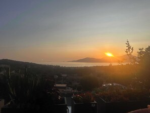Une vue sur le coucher de soleil depuis un balcon donnant sur une étendue d’eau et sur des collines lointaines.