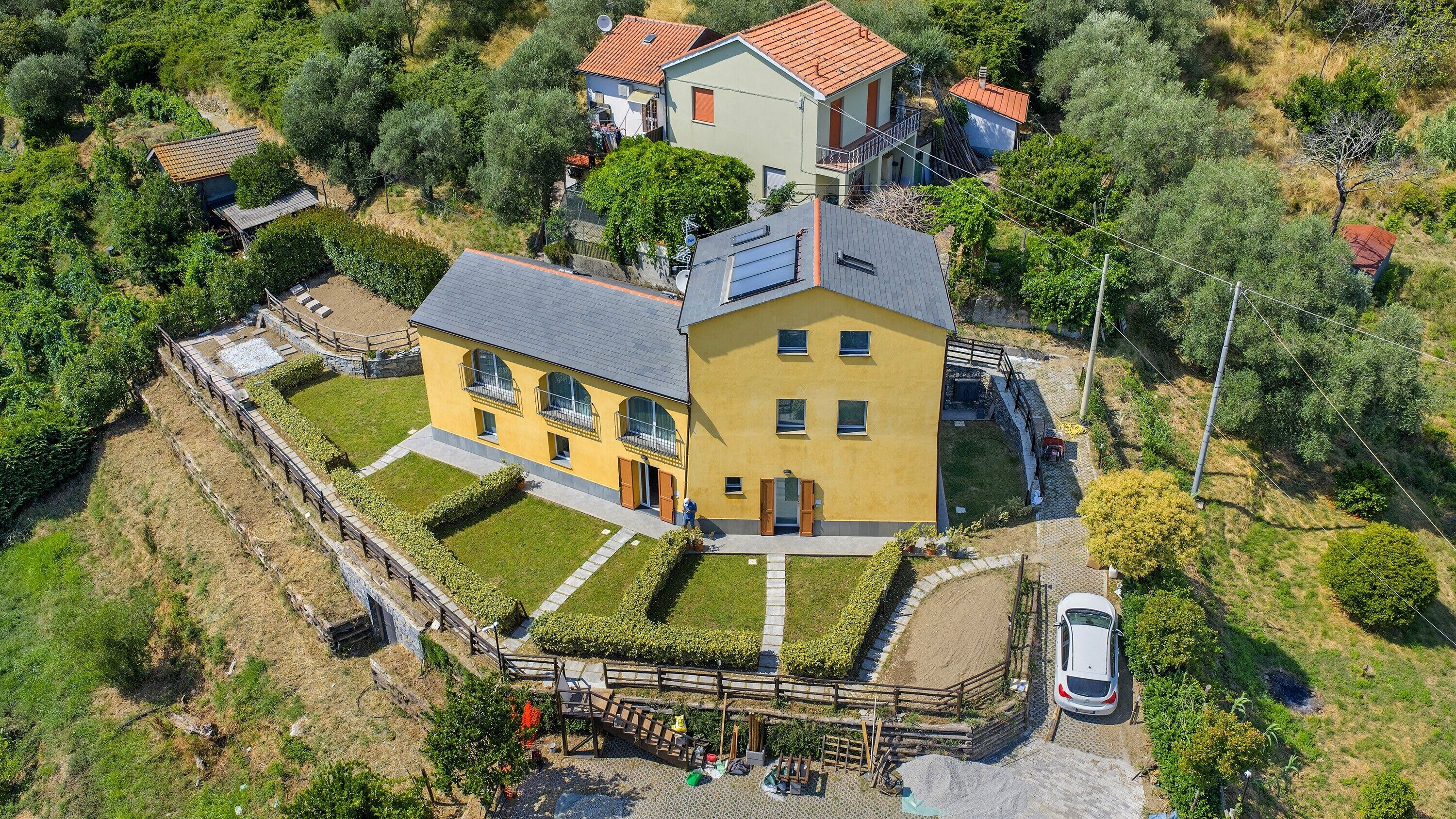A large yellow house with a grey roof, surrounded by greenery and a paved driveway.