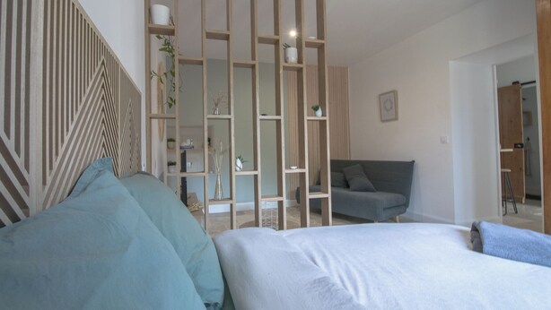 Un dormitorio con cama, un sofá gris, un panel de madera con estantes y una pared con diseño geométrico.