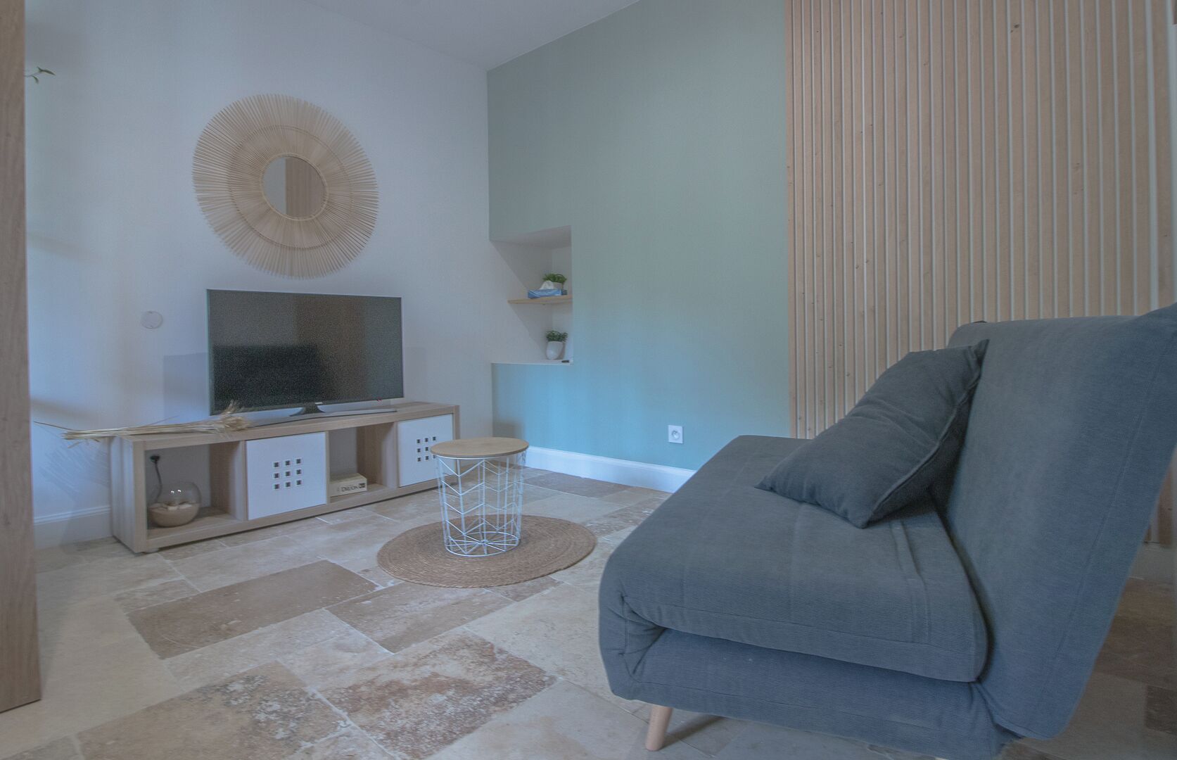 A living room with a grey sofa, a TV on a wooden stand, a woven stool, and a decorative wall piece.