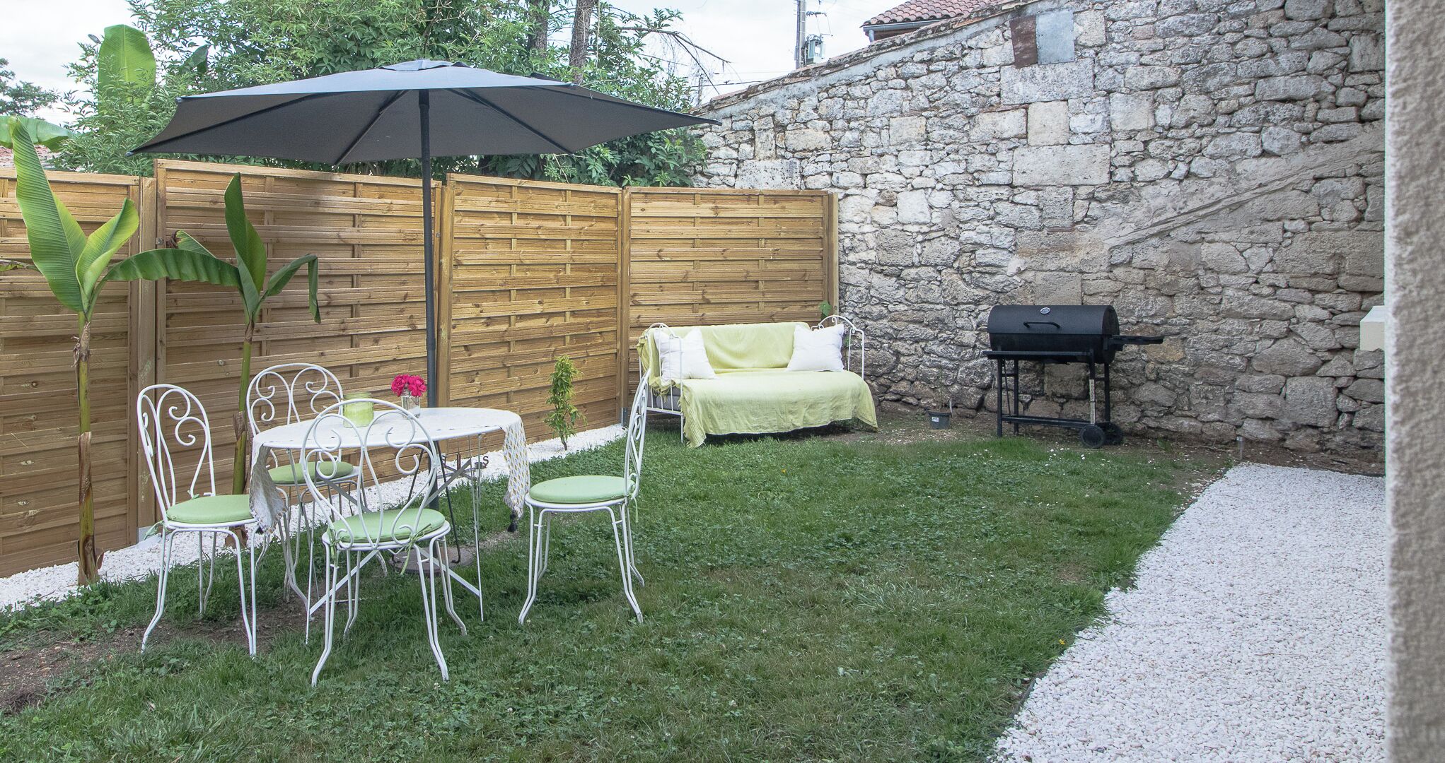 Een terras met een tafel, stoelen, een houten schutting, een stenen muur en een barbecue.