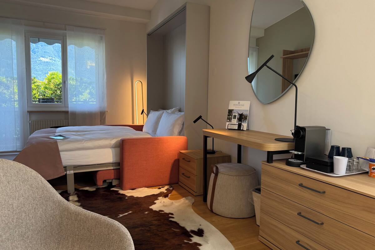 Une chambre d’hôtel équipée d’un lit, d’un bureau, d’un miroir, d’une lampe, d’une machine à café et offrant une vue sur la verdure environnante.