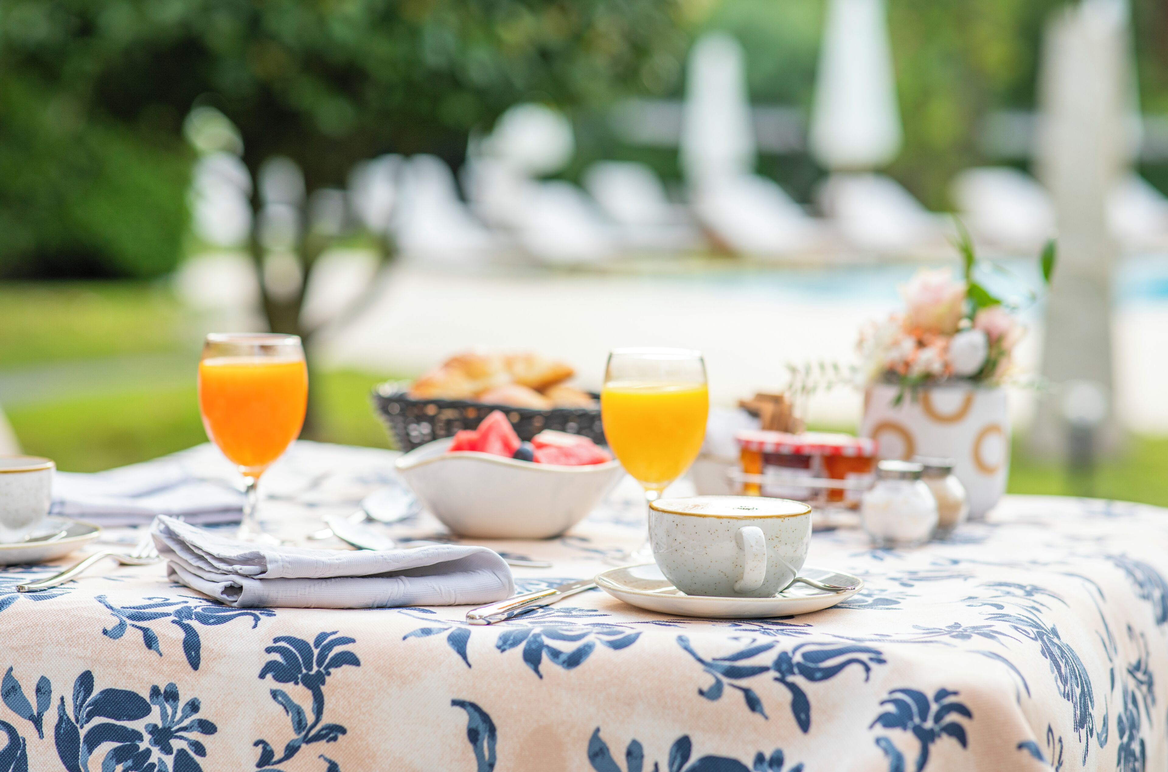 A breakfast table with a floral tablecloth, two glasses of orange juice, a bowl of fruit, and a cup of coffee.