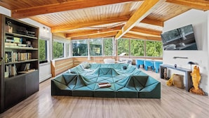 Una sala de estar moderna con un sofá seccional geométrico, vigas de madera en el techo, ventanas amplias y una estantería.