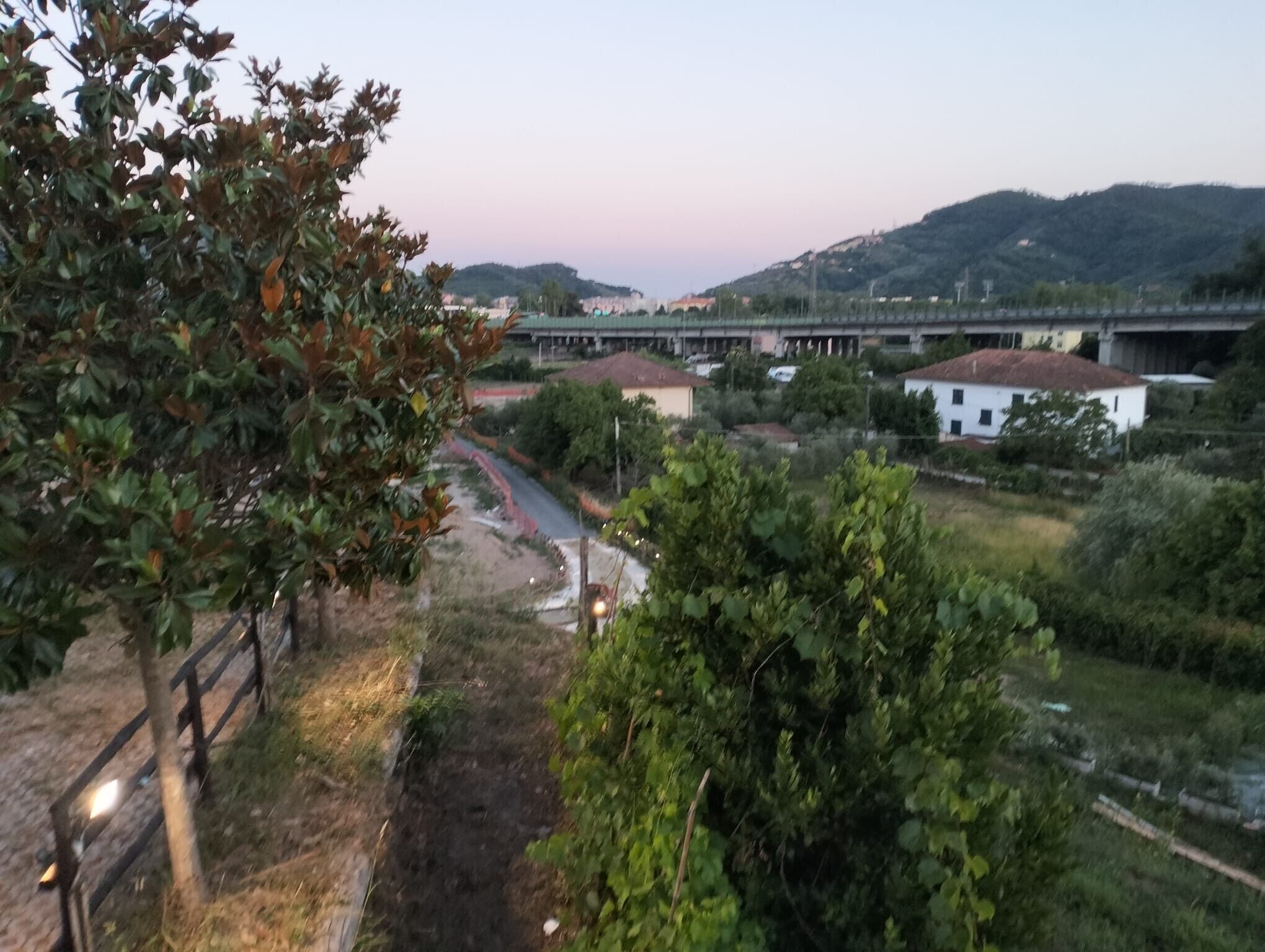 A rural landscape with a road, greenery, and a bridge in the background.
