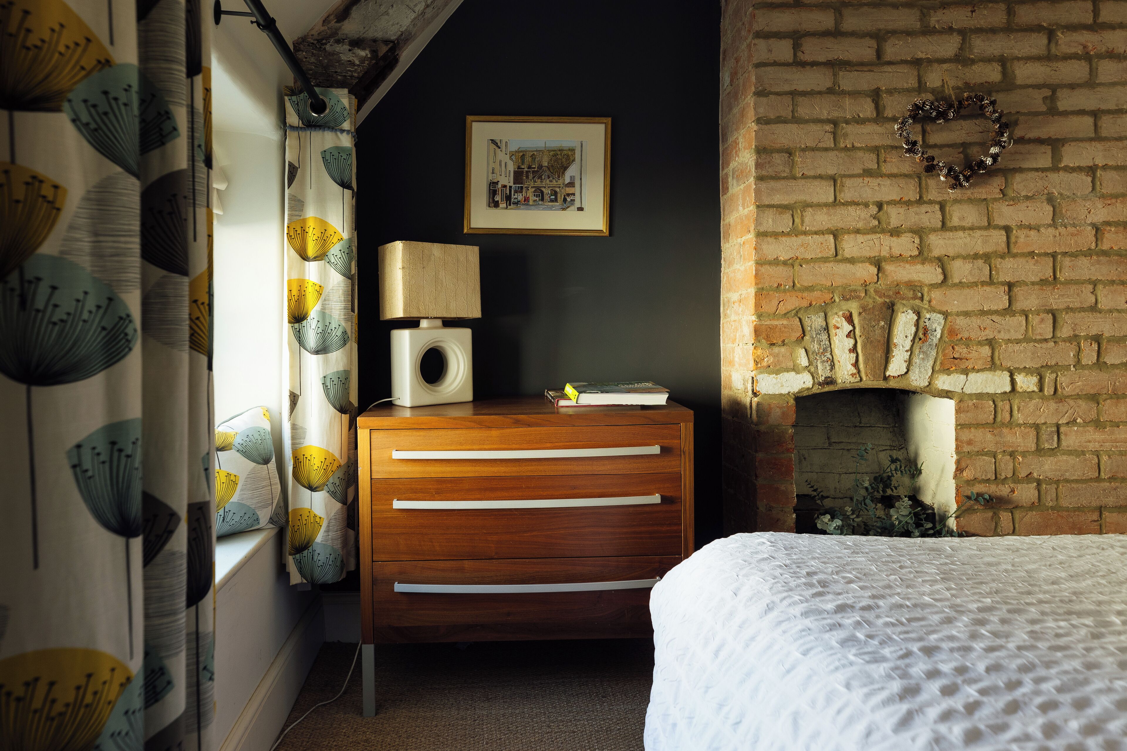 A bedroom with a wooden dresser, a white bed, a brick fireplace, and a framed picture on the wall.