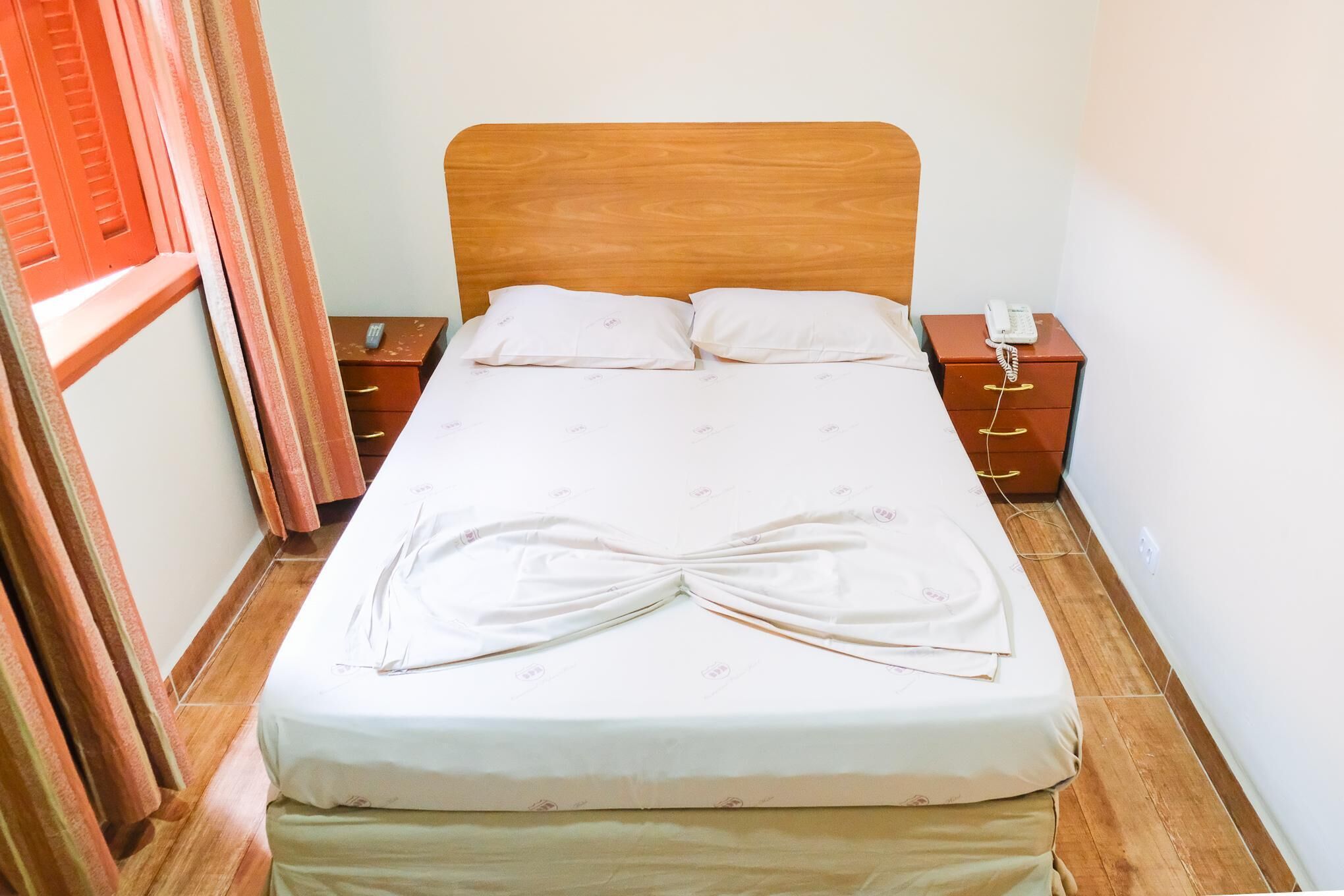 A neatly made bed with a wooden headboard, two bedside tables with phones, and a curtain partially drawn to one side.