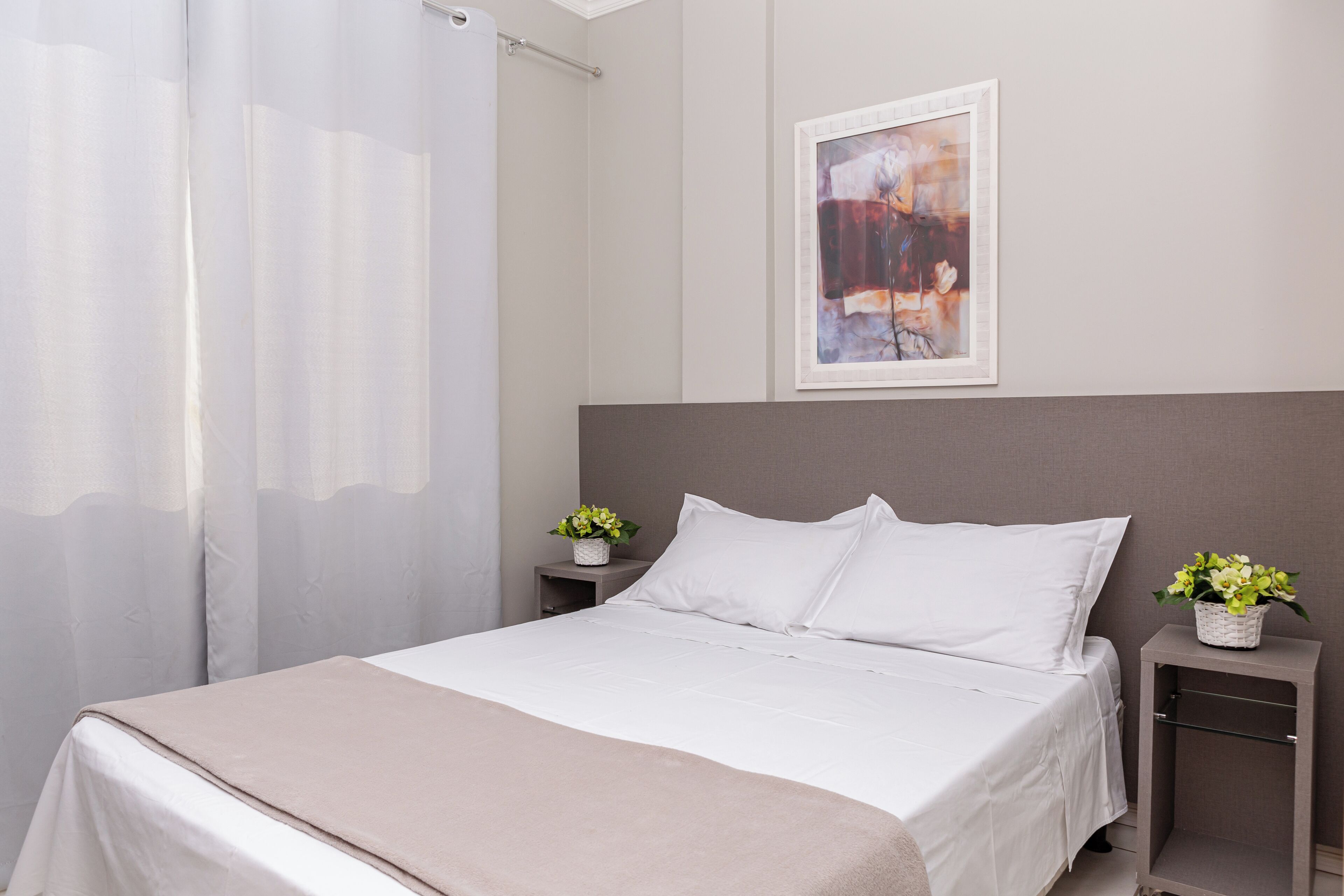 Una cama bien hecha con sábanas blancas, un cabecero gris y dos mesitas de noche con pequeños arreglos florales.