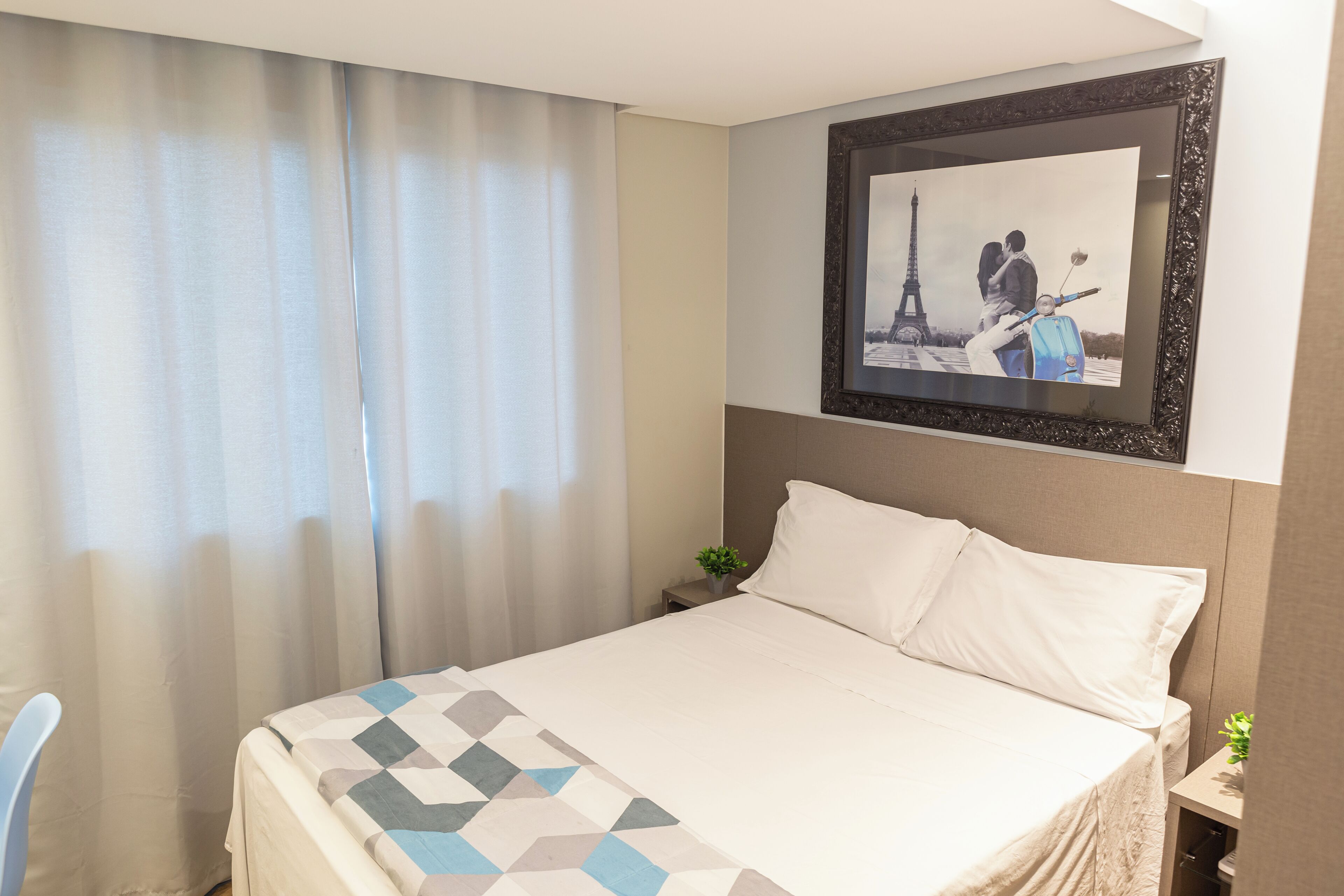 A hotel room with a bed, a framed picture of the Eiffel Tower, a small plant, and a window with curtains.