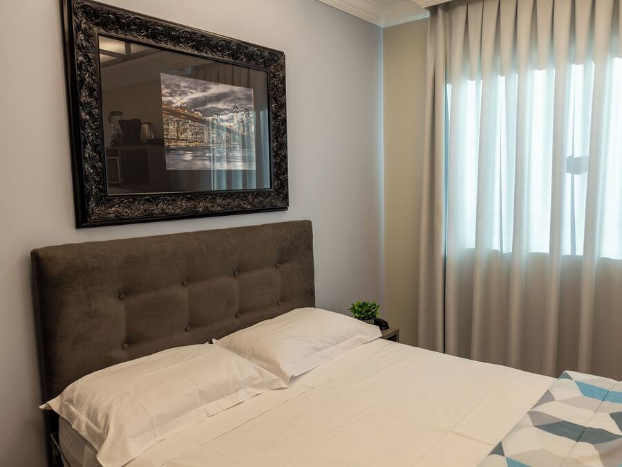 Una cama bien hecha con cabecera, un cuadro enmarcado en la pared y una ventana con cortinas.