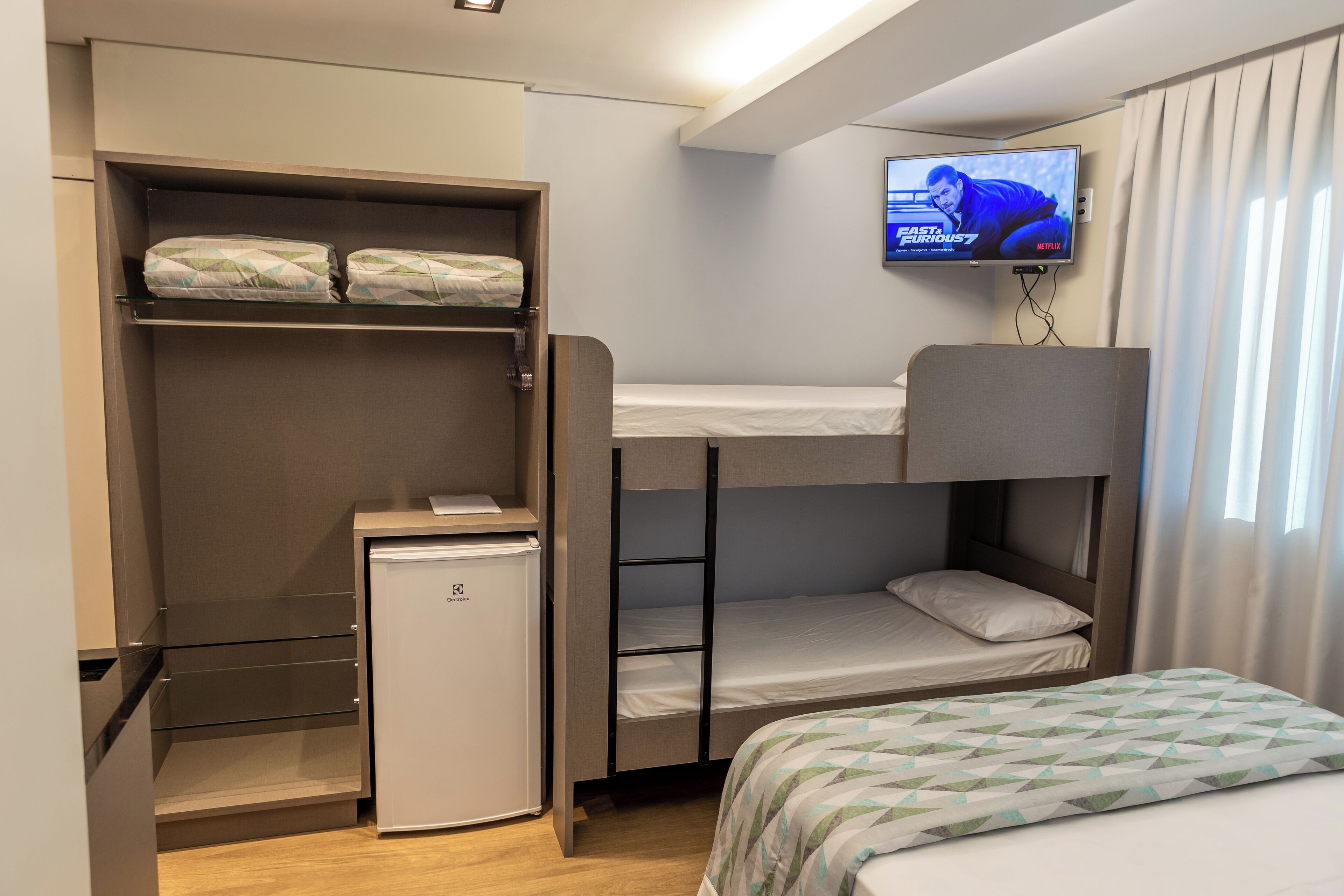 Habitación de hotel compacta con literas, un refrigerador pequeño, un televisor montado en la pared y una ventana con cortinas.