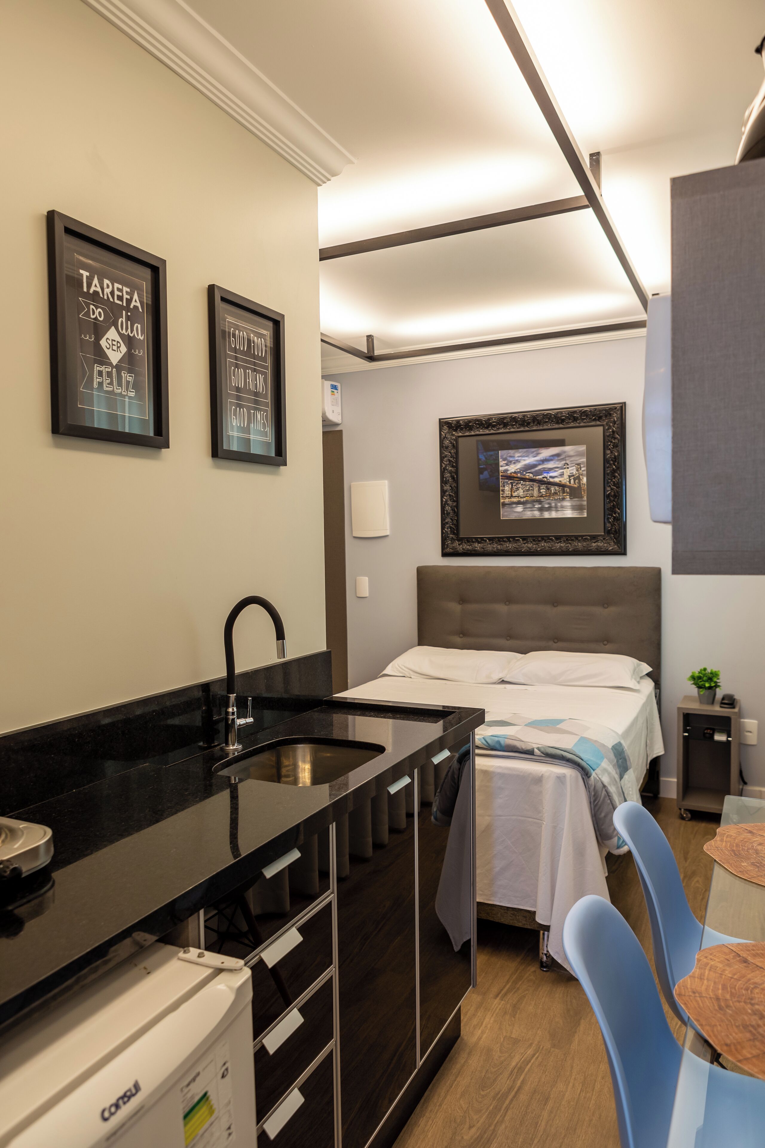 Quarto de hotel pequeno com uma cama, uma secretária com pia, uma cadeira e dois cartazes motivacionais emoldurados na parede.