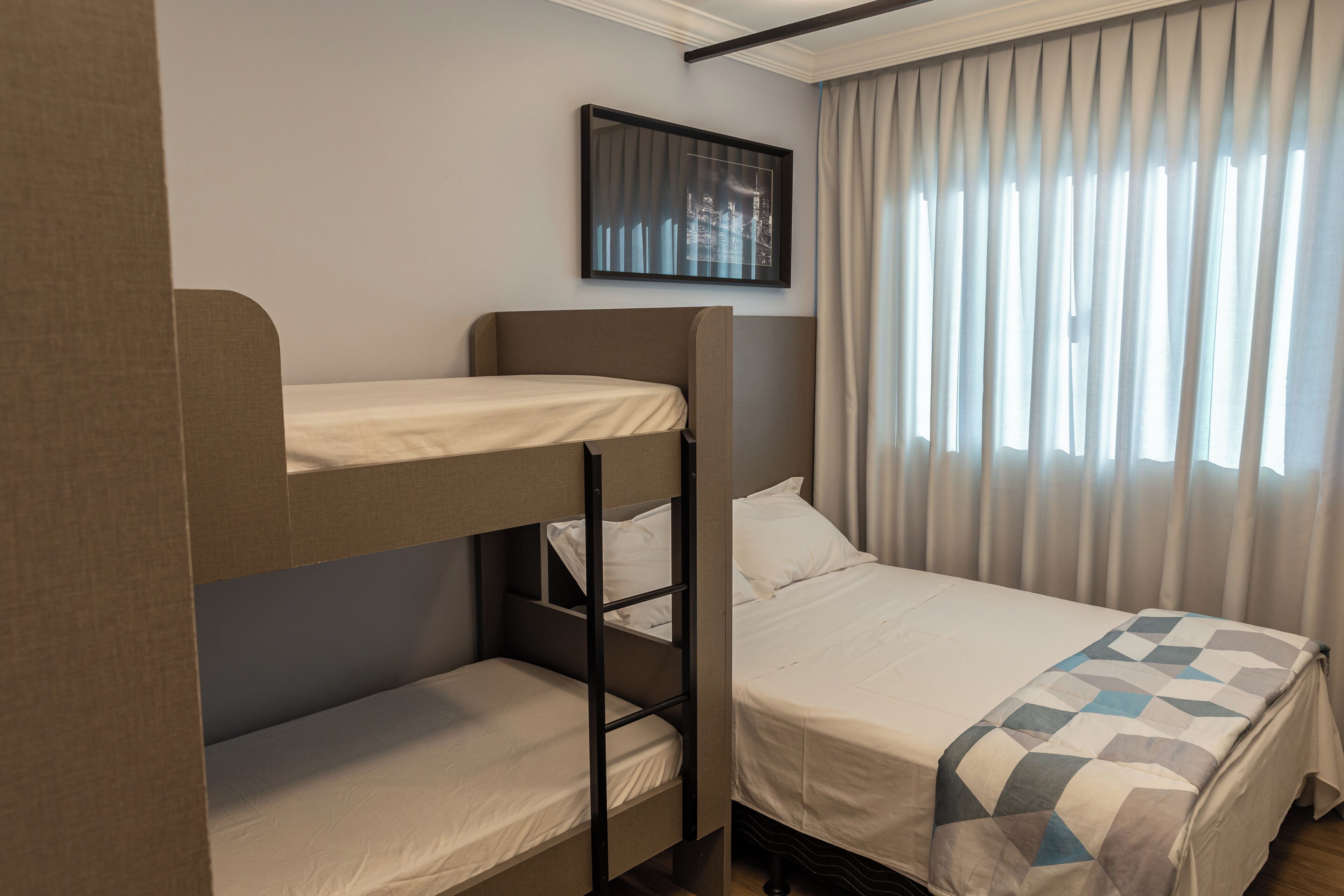A bunk bed room with a cityscape picture on the wall, a window with curtains, and a bed with a checkered blanket.