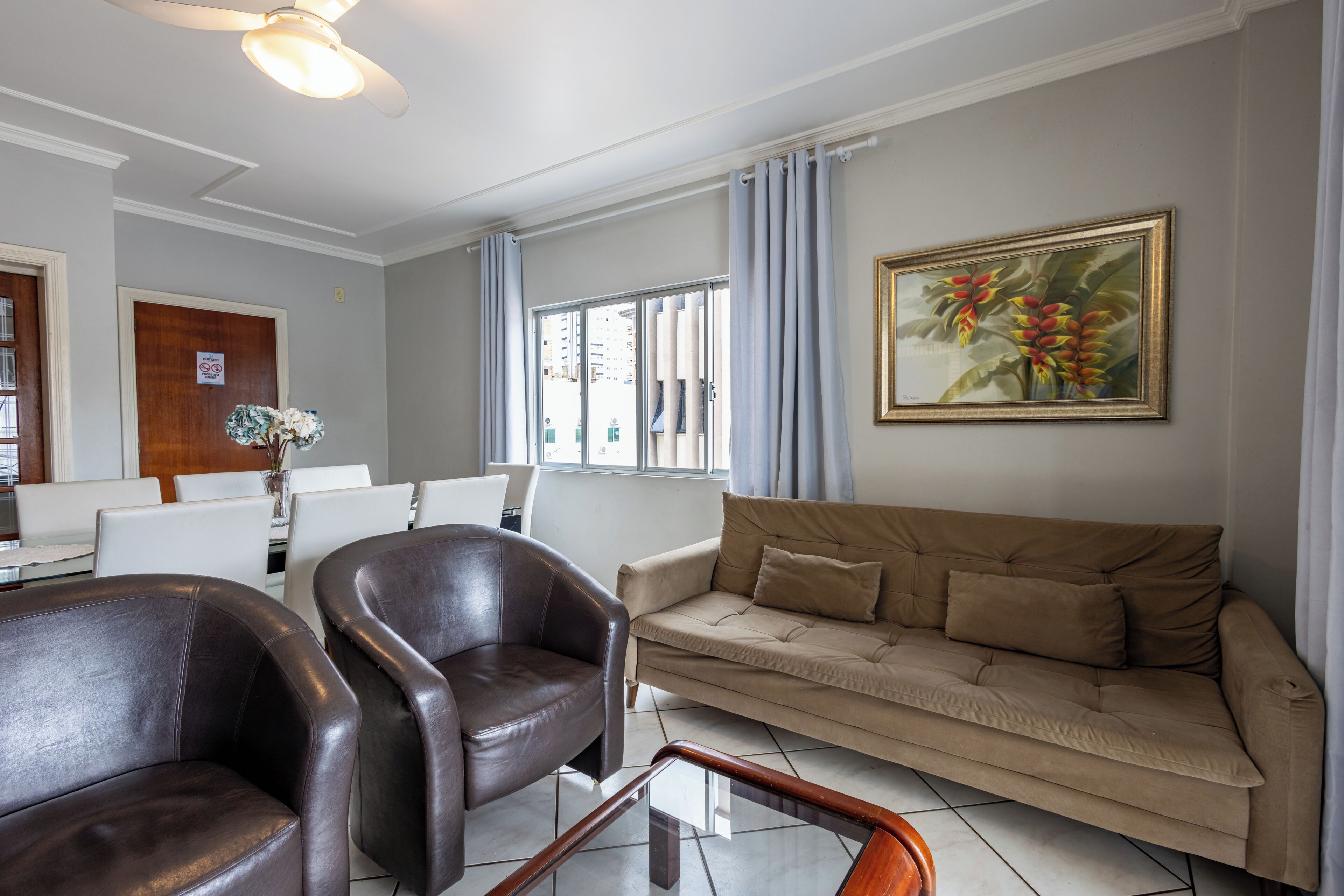 A living room with a brown leather sofa, a brown leather armchair, a glass coffee table, a dining area with white chairs, and a window with curtains.