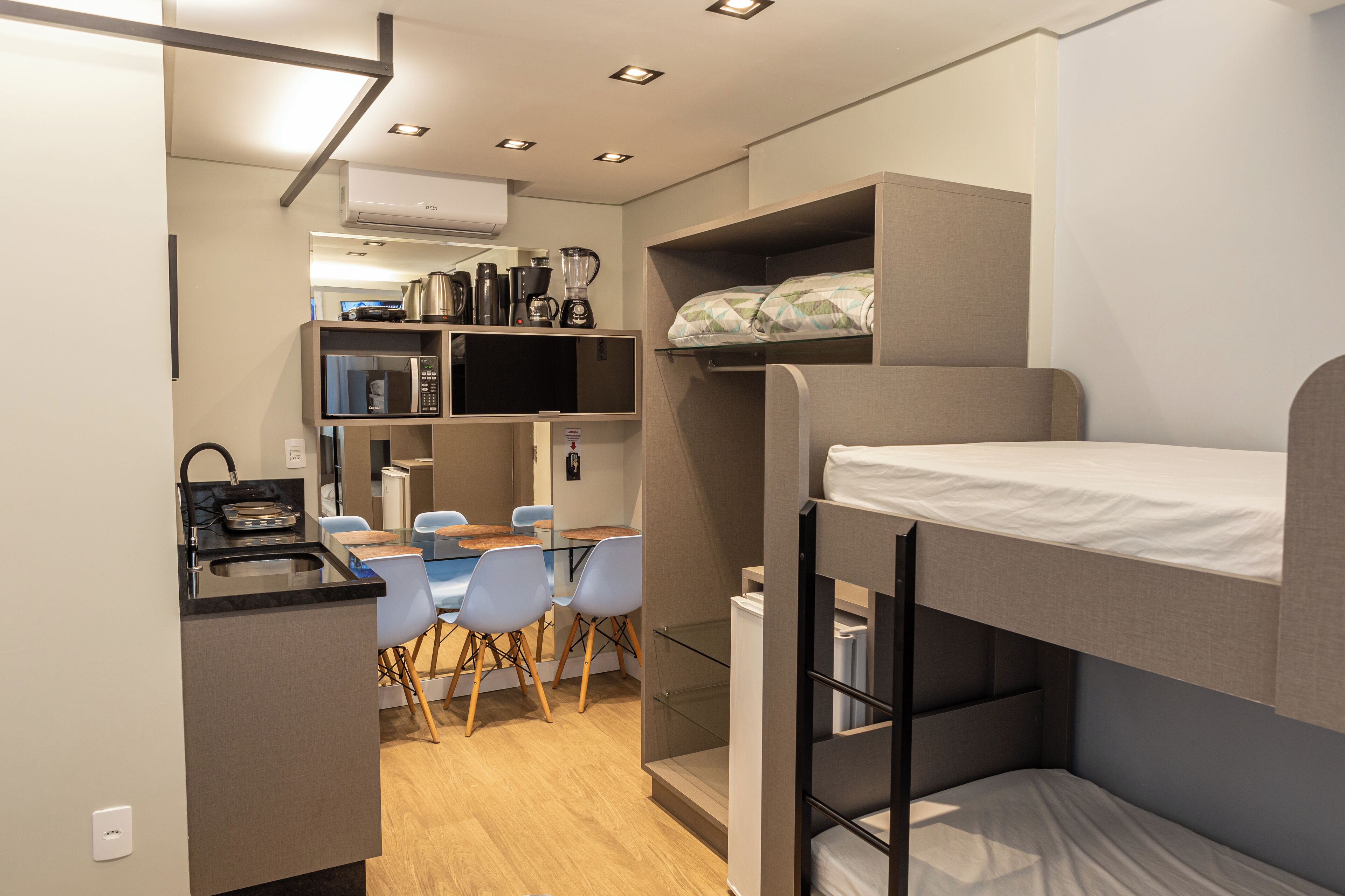 Une chambre compacte avec un lit superposé, une kitchenette équipée d’un four à micro-ondes et d’une machine à café, un petit coin repas avec des chaises et un téléviseur.
