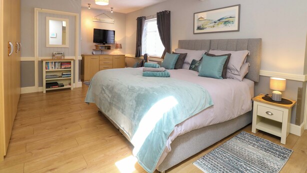 A bedroom with a bed, bedside tables, a TV, a bookshelf, and a painting on the wall. - Rainbow's End (Dereham)