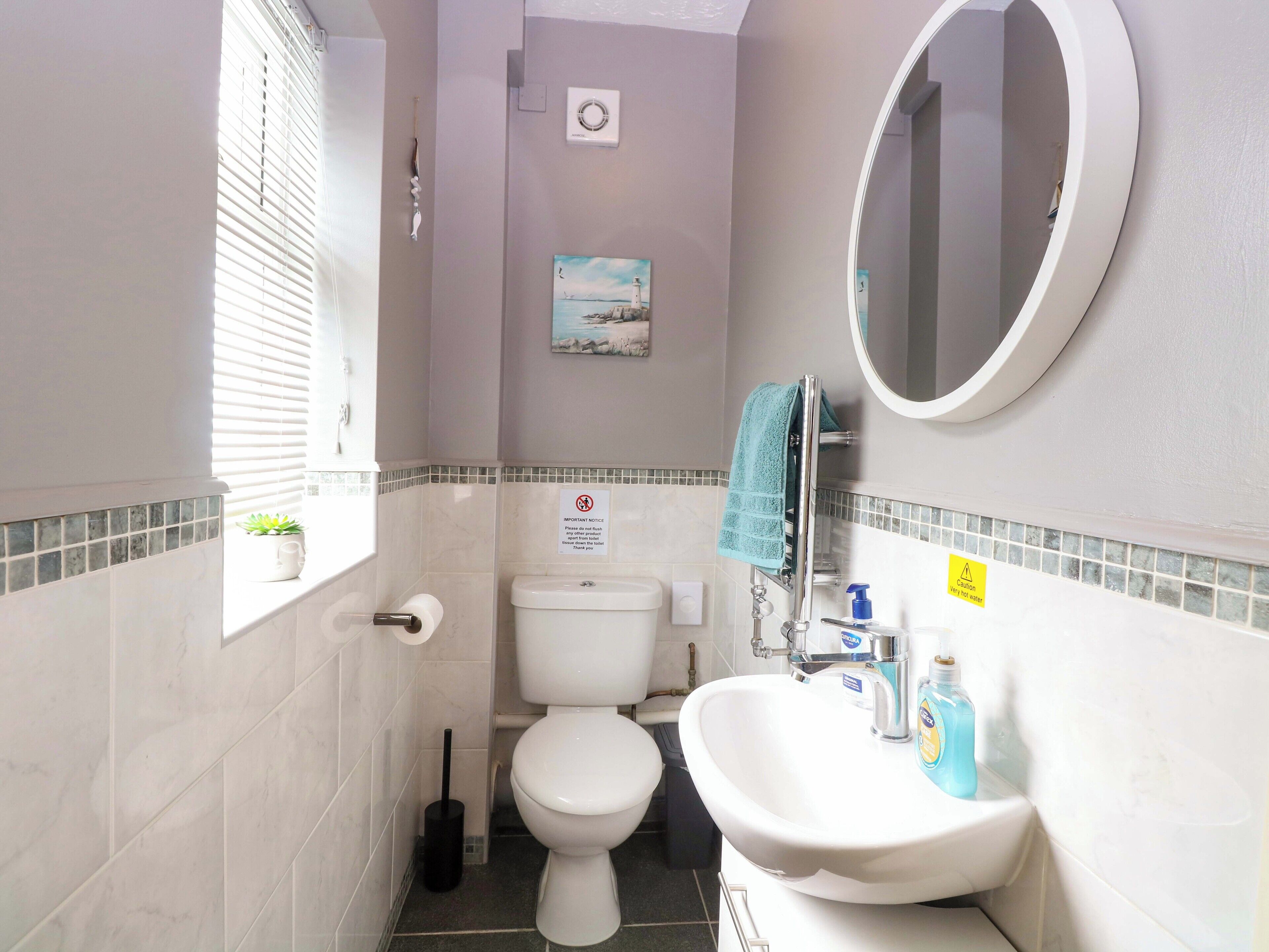 Une salle de bain avec des toilettes, un lavabo et un miroir. Il y a une fenêtre avec des stores, une petite plante et un tableau accroché au mur.