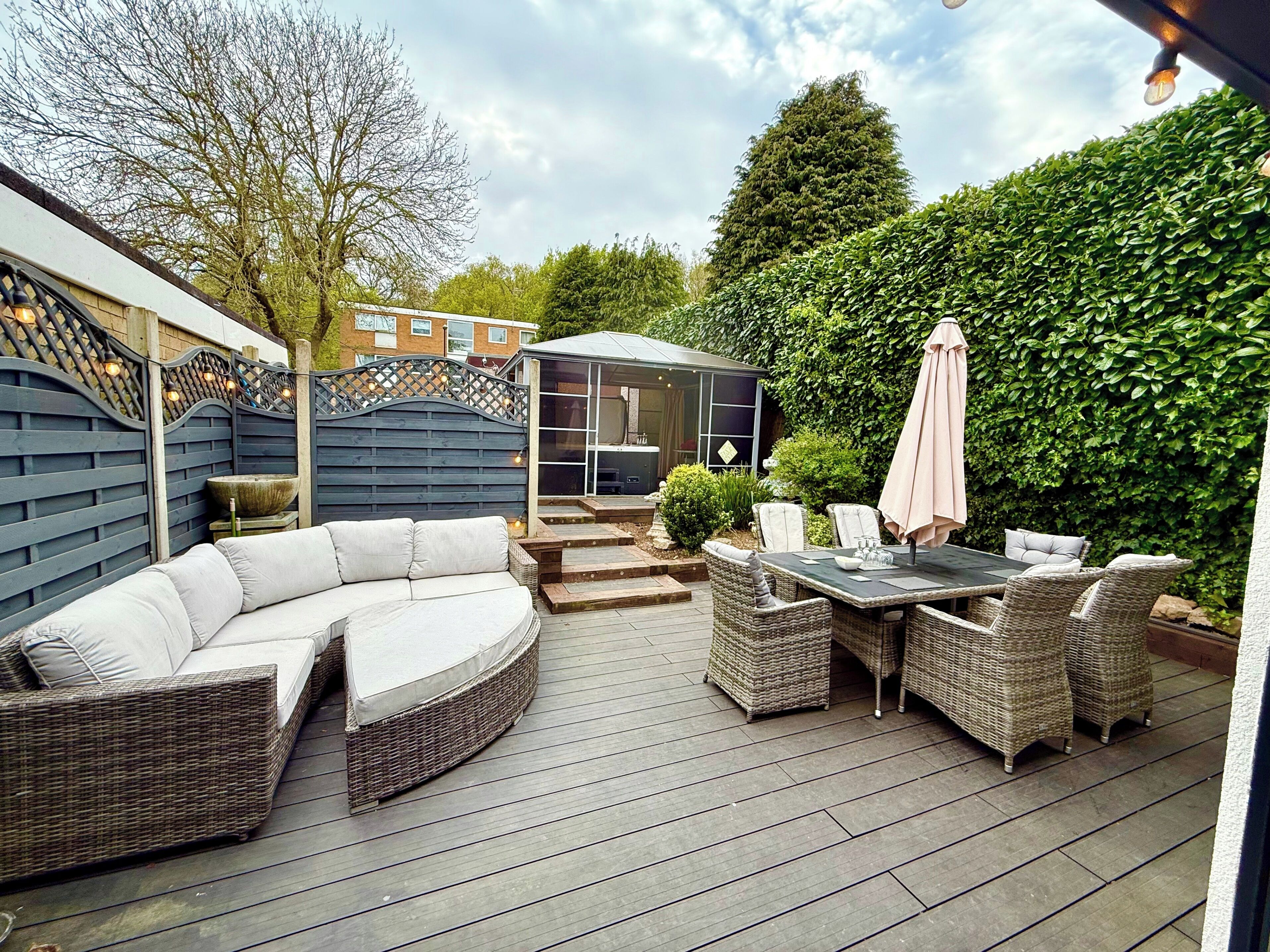 A patio with a curved sofa, wicker chairs, and a table with an umbrella. There is a small shed and a hedge in the background.