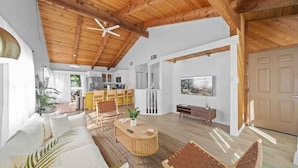 A spacious living area with a high wooden ceiling, a sofa, a coffee table, and a dining table with chairs. There is a kitchen in the background and a door leading outside.