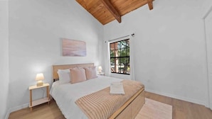 A bedroom with a bed, bedside table, lamp, and a window with a view of greenery.