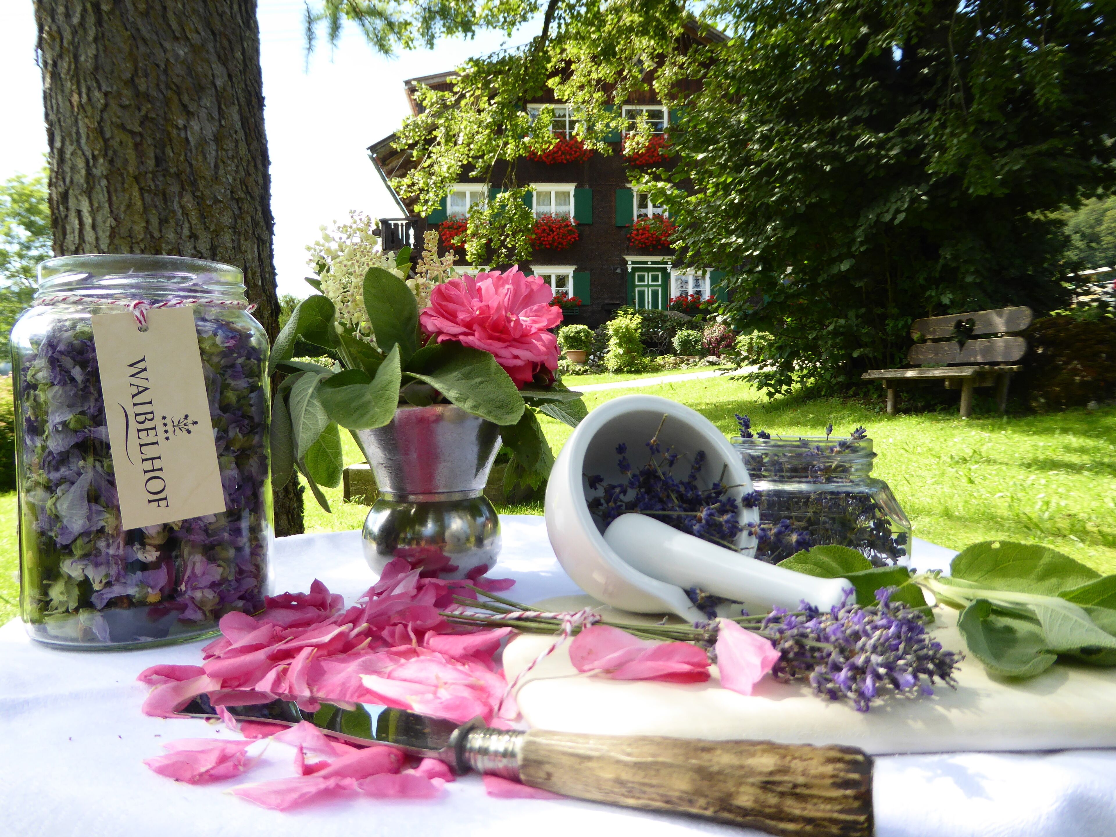 A table with a jar labeled "WABELHOF," a flower pot with pink flowers, a mortar and pestle, and scattered flower petals.