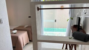 A spa room with two massage tables, a window with a view of a pool, and a wooden bench with towels.