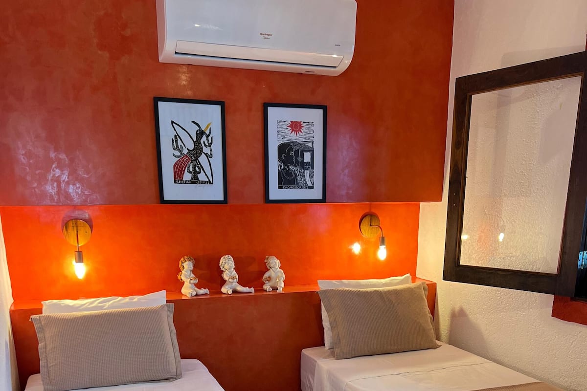 Habitación de hotel con dos camas, cada una con cubrecama y almohadas color beige, una pequeña planta decorativa y una toalla enrollada sobre la cama. Se observan lámparas de pared, un cuadro enmarcado y un equipo de aire acondicionado sobre las camas.